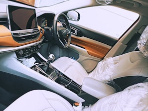 An interior view of a modern car featuring a sleek dashboard with a large touchscreen display. The steering wheel has multiple controls and the seats are covered with protective plastic wrap. The color scheme includes tones of white, brown, and black.