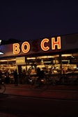 A dimly lit, cozy bistro with a large, illuminated sign reading 'BOSCH' above it. Warm, yellow lights illuminate the interior where people gather around tables, visible through the large windows. There are a few empty chairs outside the bistro, and a blurred figure on a bicycle passes by on the street.
