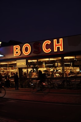A dimly lit, cozy bistro with a large, illuminated sign reading 'BOSCH' above it. Warm, yellow lights illuminate the interior where people gather around tables, visible through the large windows. There are a few empty chairs outside the bistro, and a blurred figure on a bicycle passes by on the street.