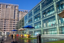 A modern urban setting with tall glass-fronted buildings reflecting sunlight. The scene includes a rooftop terrace equipped with colorful chairs and tables under blue and beige umbrellas. The area is paved and features patches of artificial grass. Both the terrace and buildings are part of a high-rise complex, depicted under a clear blue sky.