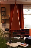 An interior room featuring modern decor elements includes a large, conical pendant lamp in the center. Behind the lamp, there are deep orange curtains and a window. A black chair is positioned near a set of shelves, holding items such as glassware and a vase. The foreground has a coffee table with decorative vases and a tray. The room's color scheme combines warm and earthy tones.