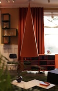 An interior room featuring modern decor elements includes a large, conical pendant lamp in the center. Behind the lamp, there are deep orange curtains and a window. A black chair is positioned near a set of shelves, holding items such as glassware and a vase. The foreground has a coffee table with decorative vases and a tray. The room's color scheme combines warm and earthy tones.