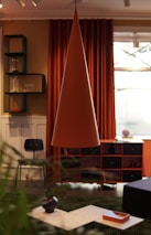 An interior room featuring modern decor elements includes a large, conical pendant lamp in the center. Behind the lamp, there are deep orange curtains and a window. A black chair is positioned near a set of shelves, holding items such as glassware and a vase. The foreground has a coffee table with decorative vases and a tray. The room's color scheme combines warm and earthy tones.
