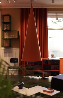 An interior room featuring modern decor elements includes a large, conical pendant lamp in the center. Behind the lamp, there are deep orange curtains and a window. A black chair is positioned near a set of shelves, holding items such as glassware and a vase. The foreground has a coffee table with decorative vases and a tray. The room's color scheme combines warm and earthy tones.