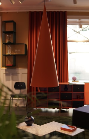 An interior room featuring modern decor elements includes a large, conical pendant lamp in the center. Behind the lamp, there are deep orange curtains and a window. A black chair is positioned near a set of shelves, holding items such as glassware and a vase. The foreground has a coffee table with decorative vases and a tray. The room's color scheme combines warm and earthy tones.