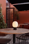 A round wooden table is situated on a wooden deck, surrounded by wire-framed chairs with dark cushions. At the center of the table is a modern, orb-shaped lamp with a soft glow. The background features tall plants and a reddish-brown wall with a black-framed glass door.