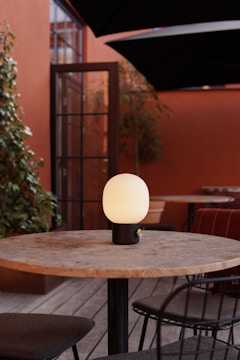 A round wooden table is situated on a wooden deck, surrounded by wire-framed chairs with dark cushions. At the center of the table is a modern, orb-shaped lamp with a soft glow. The background features tall plants and a reddish-brown wall with a black-framed glass door.