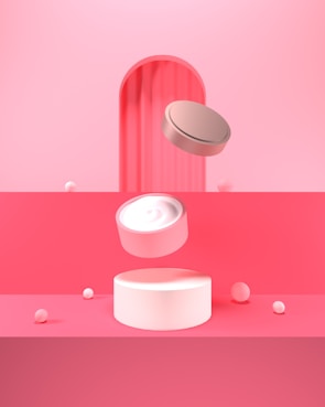 A minimalist and abstract composition featuring cylindrical objects against a pink and red backdrop. The central object appears to be a floating jar or container, possibly for cream or cosmetics, with a lid hovering above it. Small white spheres are scattered around, adding to the surreal and modern aesthetic.