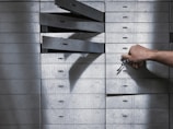 A hand holding a set of keys is reaching to open a series of safety deposit boxes. Some boxes are open, and the keyholes are visible. The boxes are metallic, numbered, and arranged in an organized manner.