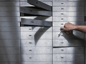 A hand holding a set of keys is reaching to open a series of safety deposit boxes. Some boxes are open, and the keyholes are visible. The boxes are metallic, numbered, and arranged in an organized manner.