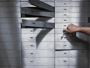 A hand holding a set of keys is reaching to open a series of safety deposit boxes. Some boxes are open, and the keyholes are visible. The boxes are metallic, numbered, and arranged in an organized manner.