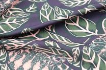 A fabric with a bold botanical pattern featuring large, stylized green leaves outlined in white against a dark background. The texture of the material is evident with slight folding, enhancing the three-dimensional appearance of the leaves. Included in the pattern are representations of small animals, adding a whimsical touch to the design.
