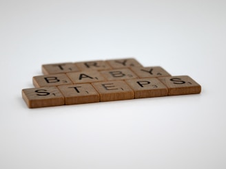 Wooden letter tiles spelling out the phrase 'TRY BABY STEPS' are arranged in a staggered line on a plain white surface. Each tile resembles those used in board games, featuring a single letter and a point value in the corner.