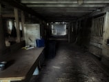 A dimly lit, rustic barn interior with wooden beams and walls. The space is cluttered with various items, including a table on the left holding tools and supplies. The floor appears dusty and covered in straw, with animal stalls lining the right side. Light filters through windows on the left, creating a contrast with the dark, shadowy corners.
