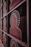 Metallic panels are designed with cut-out silhouettes of seated figures, likely representing a meditative or spiritual theme. Each silhouette is encircled by a radiating pattern, resembling rays, and arranged in a grid-like structure. The surface shows a weathered, rusty finish with a vintage aesthetic.