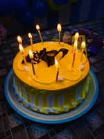 A round cake with bright yellow frosting is adorned with nine lit candles and chocolate decorations on top. The cake is placed on a blue-rimmed plate on a patterned tablecloth. In the background, there are various packaged snacks and partially visible blue balloons.