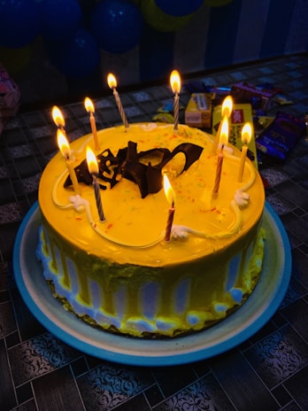 A round cake with bright yellow frosting is adorned with nine lit candles and chocolate decorations on top. The cake is placed on a blue-rimmed plate on a patterned tablecloth. In the background, there are various packaged snacks and partially visible blue balloons.