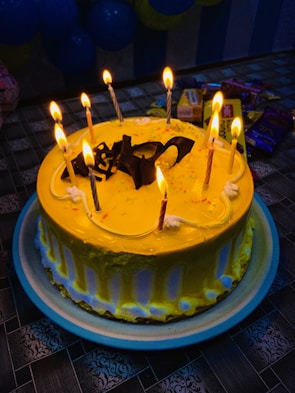 A round cake with bright yellow frosting is adorned with nine lit candles and chocolate decorations on top. The cake is placed on a blue-rimmed plate on a patterned tablecloth. In the background, there are various packaged snacks and partially visible blue balloons.