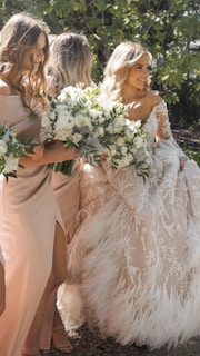 A bride in an intricately detailed white gown with feather and lace embellishments stands with her bridesmaids, who are dressed in light pink dresses. They hold lush, white floral bouquets with greenery. The setting is outdoors with sunlight filtering through the trees, creating a warm and inviting atmosphere.