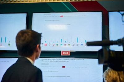 Three large screens display election results in a control room environment. The screens show various graphs and text in Russian, indicating processed protocols and voting results for different districts. A person is facing the screens, possibly analyzing the data, while a camera or similar equipment is visible in the foreground.