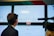 Three large screens display election results in a control room environment. The screens show various graphs and text in Russian, indicating processed protocols and voting results for different districts. A person is facing the screens, possibly analyzing the data, while a camera or similar equipment is visible in the foreground.