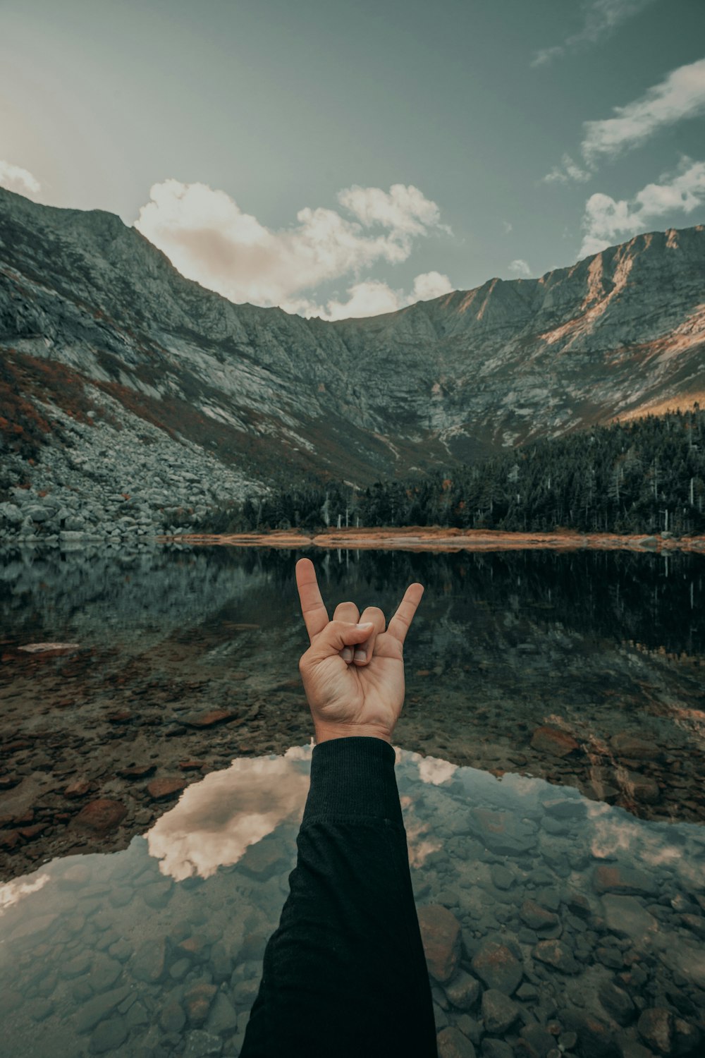 a person making a peace sign in front of a mountain lake