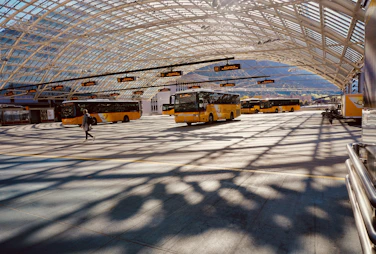 A large indoor bus terminal with multiple yellow buses parked under a high, glass-domed ceiling. A person carrying a bag walks across the terminal floor, which is patterned with shadows from the architectural framework. Mountains can be seen in the background through the transparent ceiling and walls.