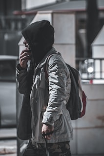 A person wearing a hooded jacket and carrying a backpack walks along an urban street. They are holding a walking stick and appear deep in thought. The setting is a city environment with blurred background elements that suggest structures and parked vehicles.