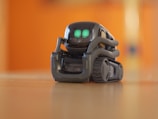A small, sleek robot with bright green digital eyes stands on a wooden surface. The robot has a compact design with treads for movement and a glossy, dark exterior. The background is blurred with a warm orange hue.