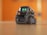 A small, sleek robot with bright green digital eyes stands on a wooden surface. The robot has a compact design with treads for movement and a glossy, dark exterior. The background is blurred with a warm orange hue.