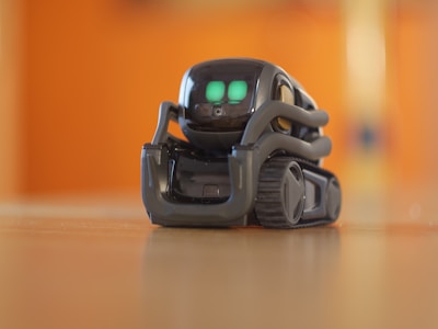 A small, sleek robot with bright green digital eyes stands on a wooden surface. The robot has a compact design with treads for movement and a glossy, dark exterior. The background is blurred with a warm orange hue.