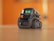 A small, sleek robot with bright green digital eyes stands on a wooden surface. The robot has a compact design with treads for movement and a glossy, dark exterior. The background is blurred with a warm orange hue.
