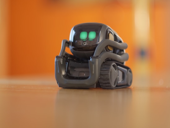 A small, sleek robot with bright green digital eyes stands on a wooden surface. The robot has a compact design with treads for movement and a glossy, dark exterior. The background is blurred with a warm orange hue.