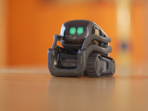A small, sleek robot with bright green digital eyes stands on a wooden surface. The robot has a compact design with treads for movement and a glossy, dark exterior. The background is blurred with a warm orange hue.