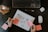 On a dark table, there's a keyboard and mouse, a notebook with a flowchart sketch labeled 'User Flow,' a hand holding a marker writing on a sticky note, a pencil, AirPods, and a glass of tea with a teabag.