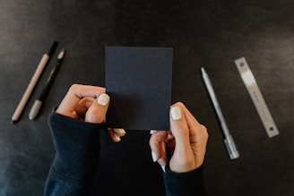 Hands with white nail polish hold a small sheet of black paper. Various writing tools, including a pencil, a pen, and a ruler, are spread out on a dark surface.