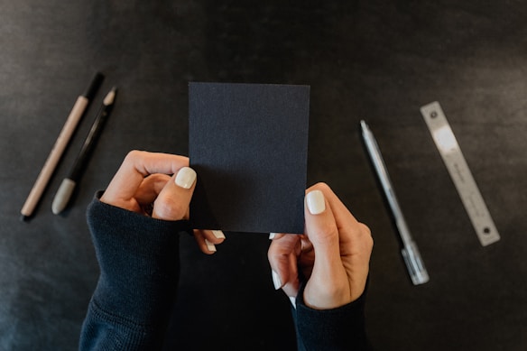Hands with white nail polish hold a small sheet of black paper. Various writing tools, including a pencil, a pen, and a ruler, are spread out on a dark surface.