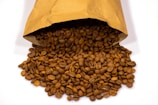 A brown paper bag is tipped over, spilling a large quantity of roasted coffee beans onto a white surface. The beans are uniformly dark brown, indicating that they have been well-roasted.