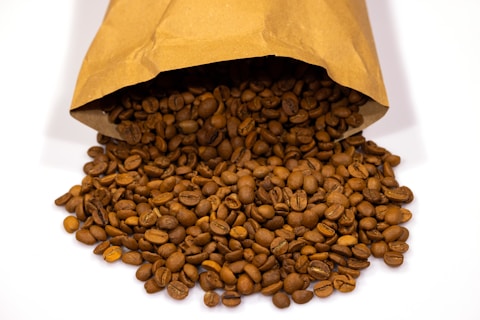 A brown paper bag is tipped over, spilling a large quantity of roasted coffee beans onto a white surface. The beans are uniformly dark brown, indicating that they have been well-roasted.