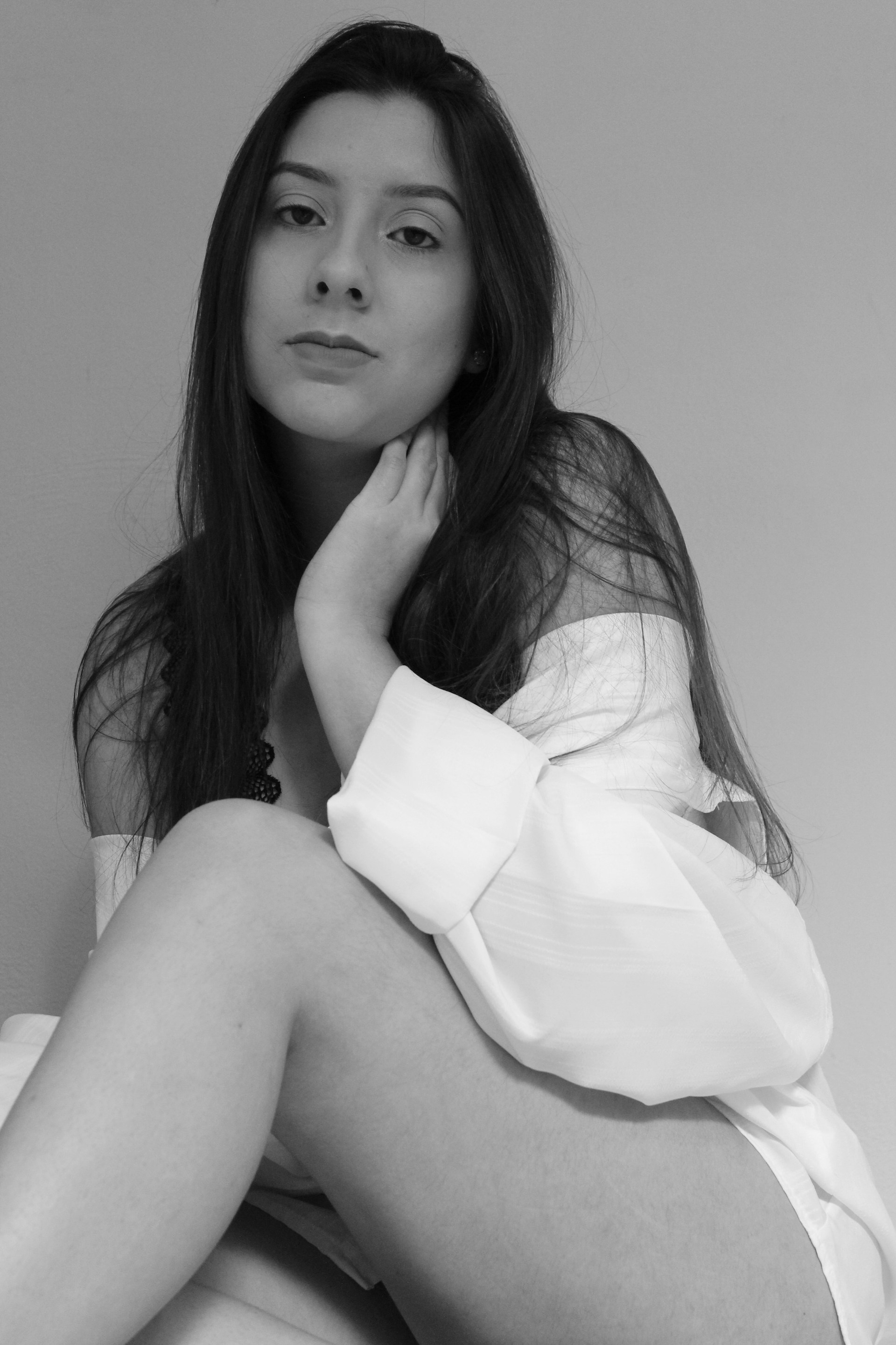 A black and white portrait of a young woman with long, dark hair. She is seated and gazes directly at the camera, wearing an off-shoulder top. One hand rests gently on her neck, adding a soft and contemplative pose.