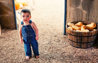 A young child wearing denim overalls stands on a brown, mulch-covered floor. To the right, there is a wooden barrel filled with squashes. In the background, pumpkins and a person are slightly visible.