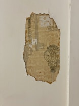A piece of old patterned wallpaper is peeling away from a white wall, revealing vintage designs that include ornate decorations and geometrical shapes. The wallpaper shows signs of aging with discoloration and rough edges.