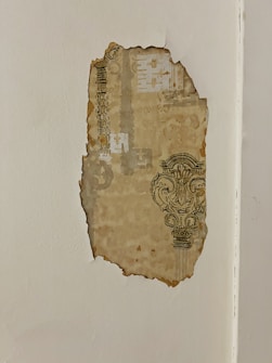 A piece of old patterned wallpaper is peeling away from a white wall, revealing vintage designs that include ornate decorations and geometrical shapes. The wallpaper shows signs of aging with discoloration and rough edges.