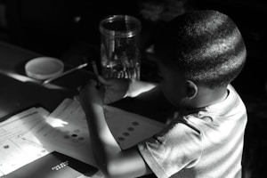 A young child sits at a table concentrating on paperwork. The child holds a pencil and appears focused on homework or activities in front of them, with shadows cast across the scene. A jar filled with writing utensils and a bowl or dish sit nearby.