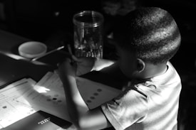 A young child sits at a table concentrating on paperwork. The child holds a pencil and appears focused on homework or activities in front of them, with shadows cast across the scene. A jar filled with writing utensils and a bowl or dish sit nearby.