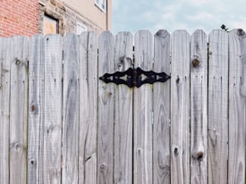 A wooden fence with a prominent black hinge, set against a background of a brick and stone building.