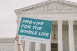 A hand holds up a sign that reads 'Pro-life for the whole life' in front of a classical government building with columns and a stone relief depicting human figures.