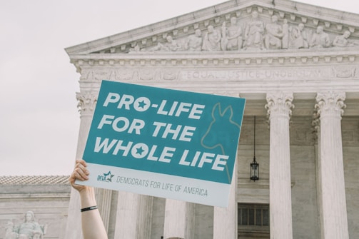 A hand holds up a sign that reads 'Pro-life for the whole life' in front of a classical government building with columns and a stone relief depicting human figures.