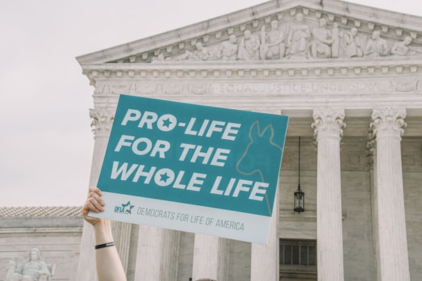 A hand holds up a sign that reads 'Pro-life for the whole life' in front of a classical government building with columns and a stone relief depicting human figures.