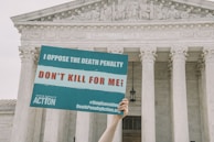 A hand is holding a protest sign in front of a large neoclassical building with columns. The sign reads 'I Oppose the Death Penalty' and 'Don't Kill for Me!', along with hashtags and a website link.